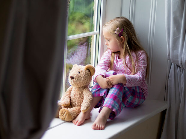 Mädchen sitzt mit Ihrem Teddy auf einem breiten Fenstersims und beide schauen hinaus