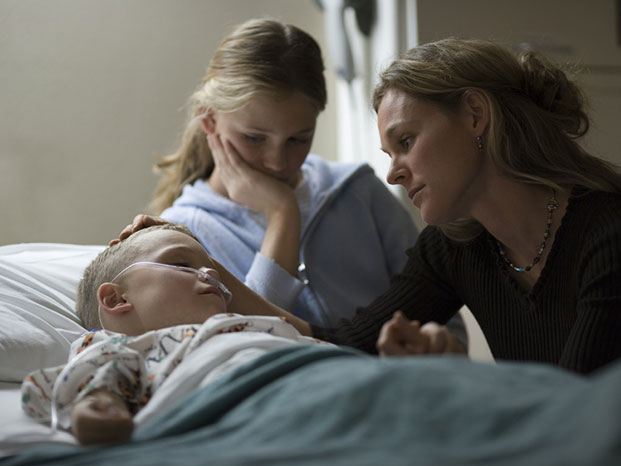 Kind im Krankenbett. Schwester und Mutter sitzen besorgt dabei.